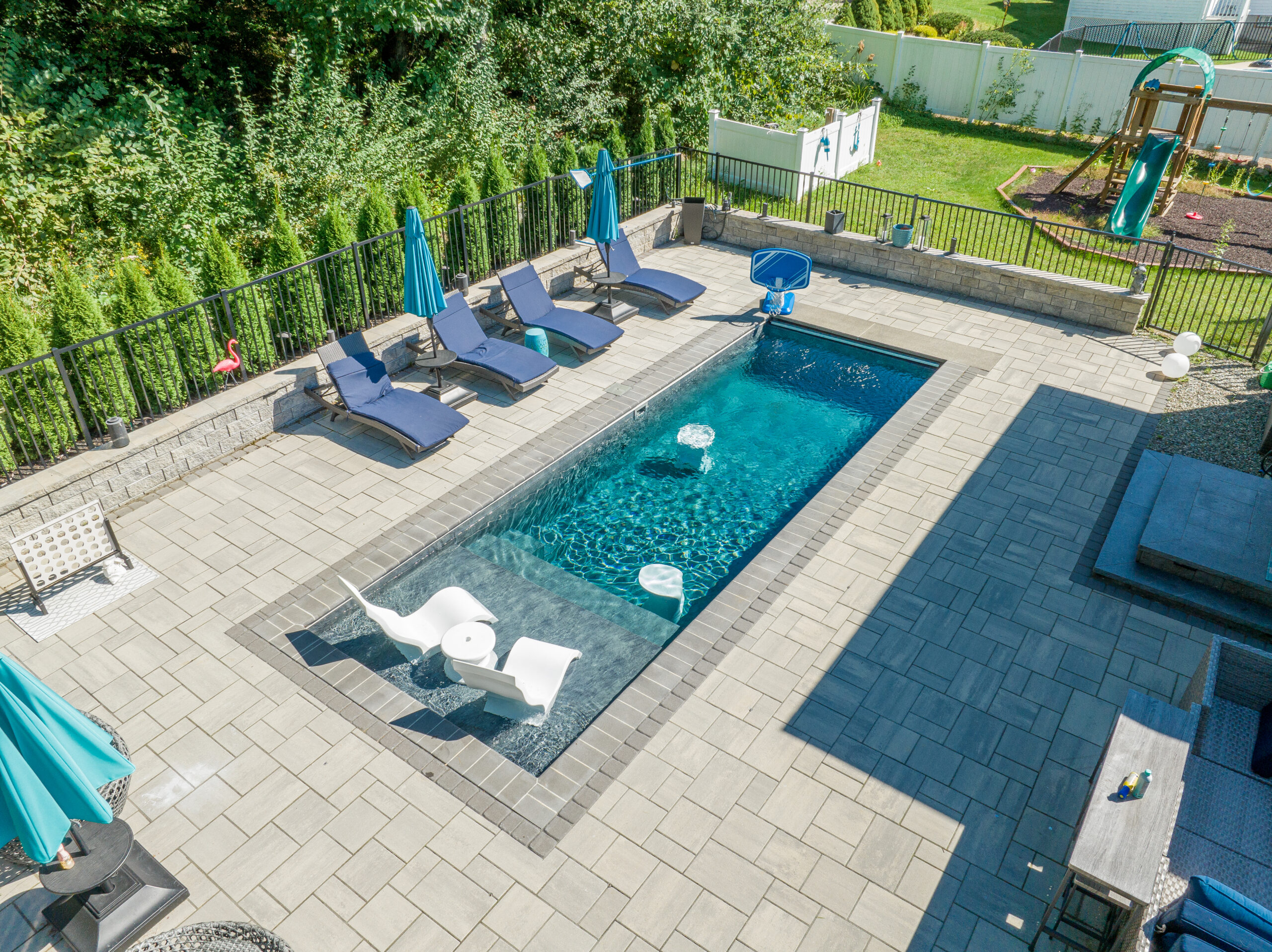 A custom plunge pool surrounded by a spacious light-gray stone patio. The pool features a clear blue tiled interior with a built-in bench and attached spa section.