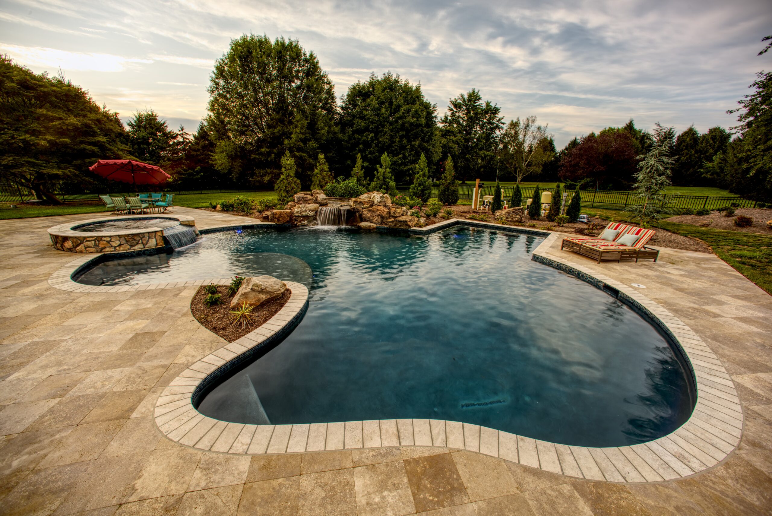 A professionally designed, custom-shaped in-ground swimming pool and surrounding patio at dusk. The pool has a dark interior and a light-colored brick coping, which contrasts with the larger, light-colored flagstone patio.