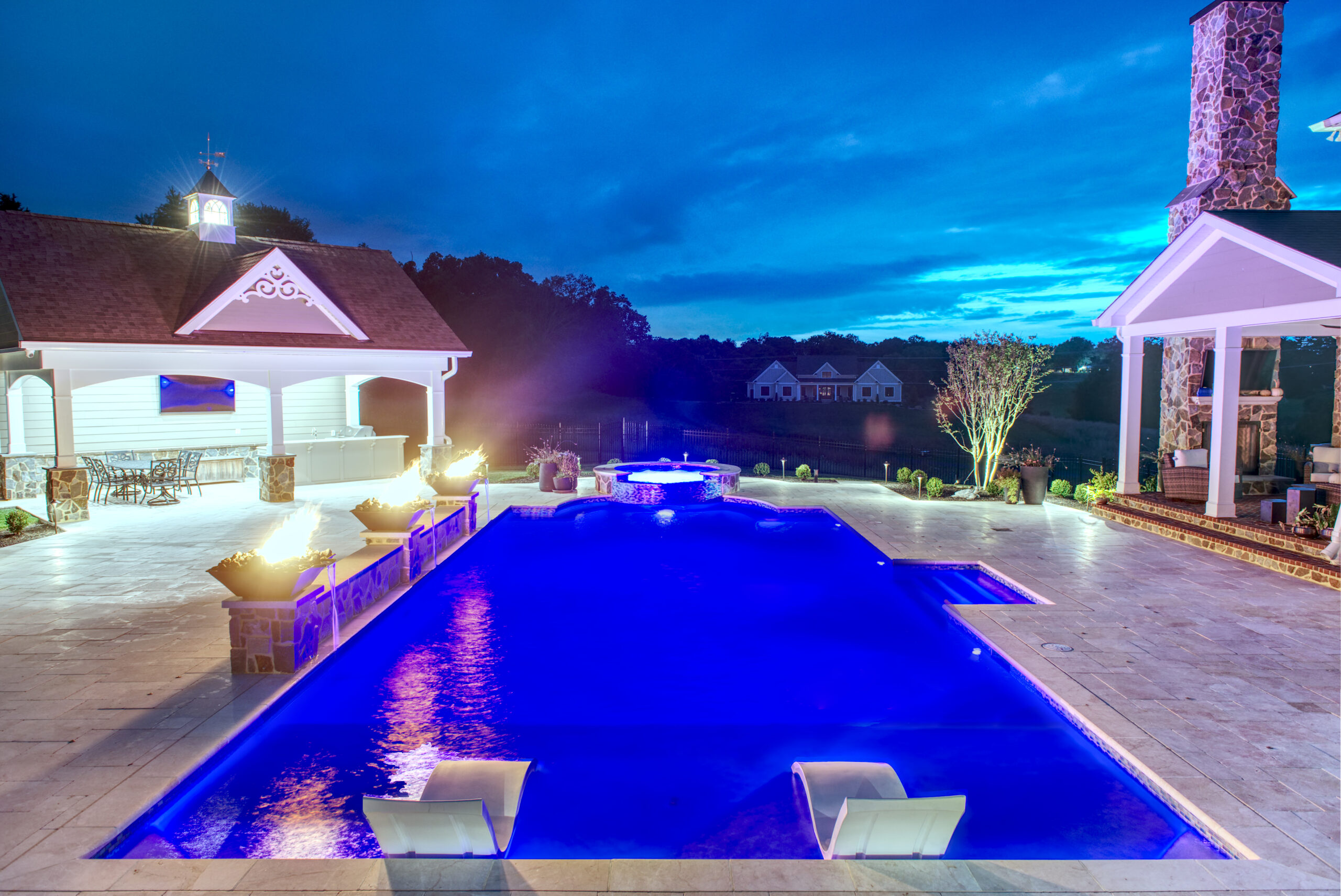 An evening photo of a luxurious rectangular swimming pool with bright blue interior lights. The pool deck is a light-colored tile, and the setting includes a structure to the left with a covered seating area and a cupola on the roof, as well as an open-sided structure to the right featuring a stone fireplace or chimney.