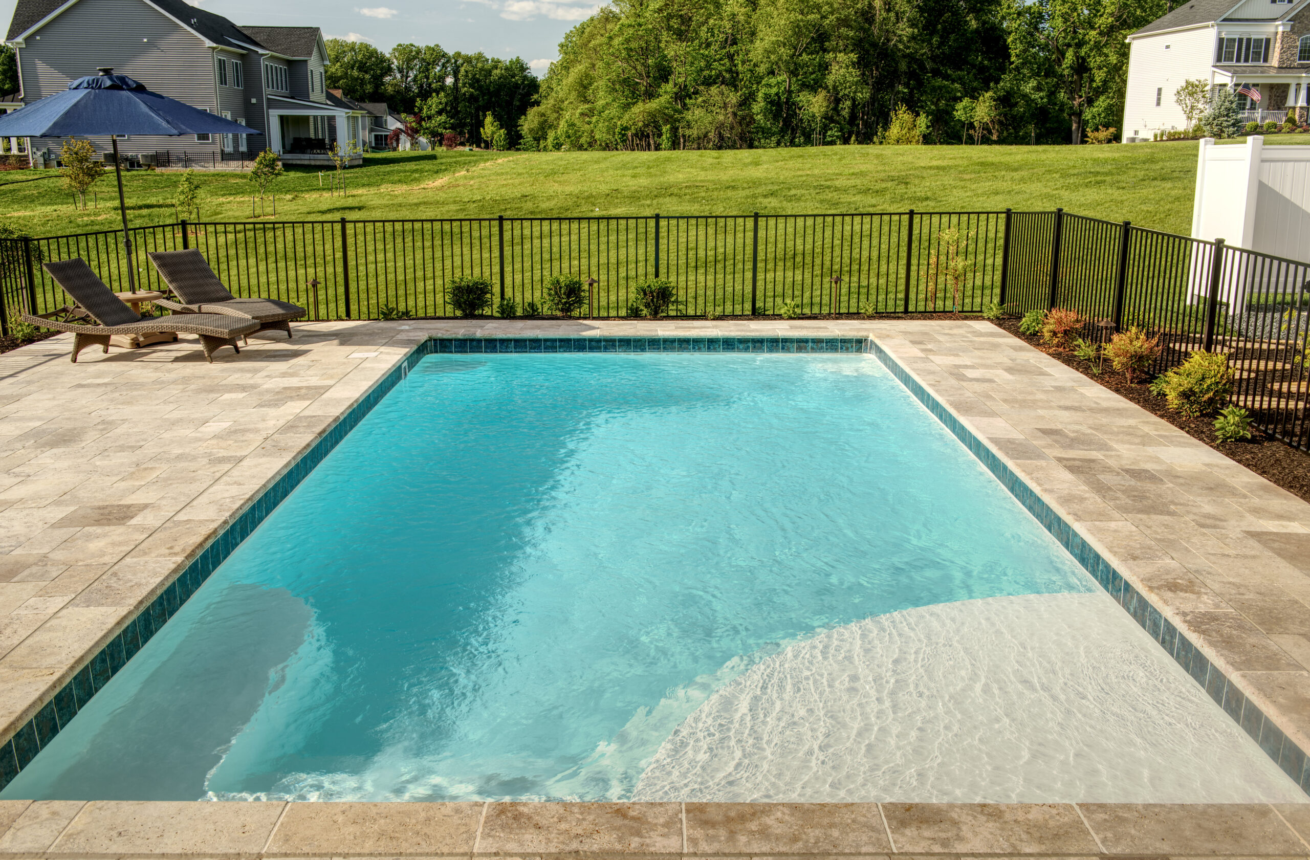 The image depicts a rectangular fiberglass in-ground swimming pool surrounded by a beige paved patio and a black metal fence.