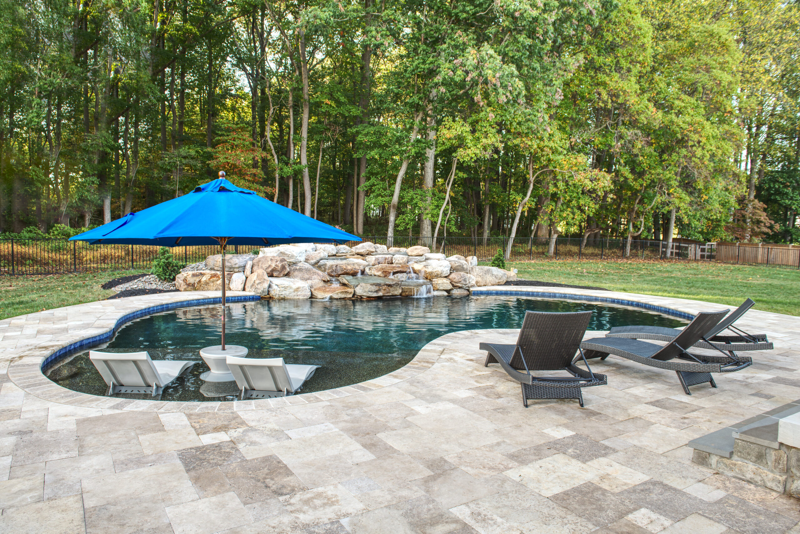 an outdoor pool area surrounded by a patio and a wooded backyard. The free-form swimming pool is paved with light-colored, patterned stone tiles. A rock waterfall feature is visible at the far edge of the pool.