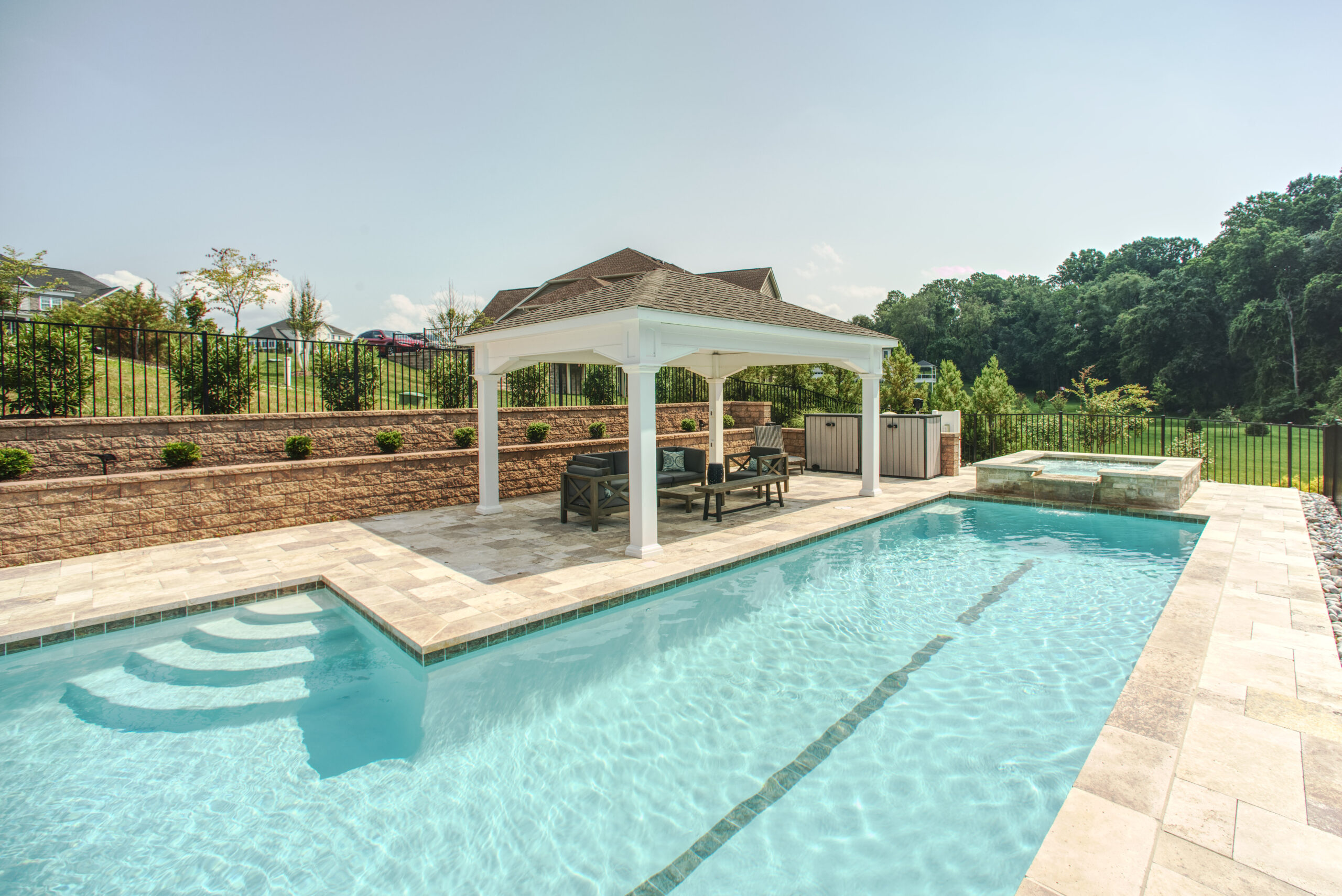 a rectangular swimming pool with light blue water and stone coping around the edge. There is a tanning ledge with built-in steps on the left side of the pool. Behind the pool, a white wooden pergola with a dark roof covers an outdoor seating area.