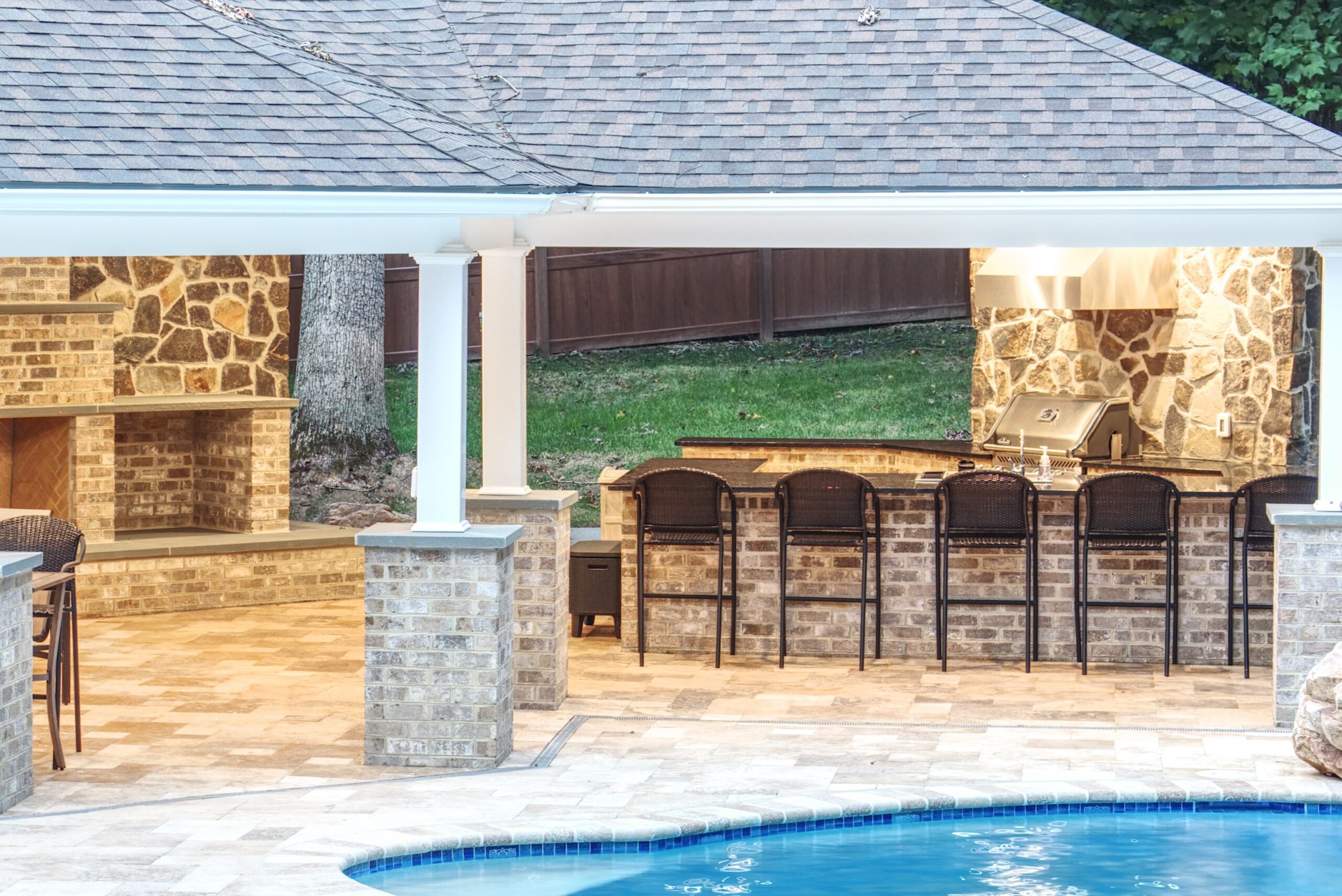 An outdoor covered patio featuring a kitchen area, fireplace, and bar seating overlooking a swimming pool.