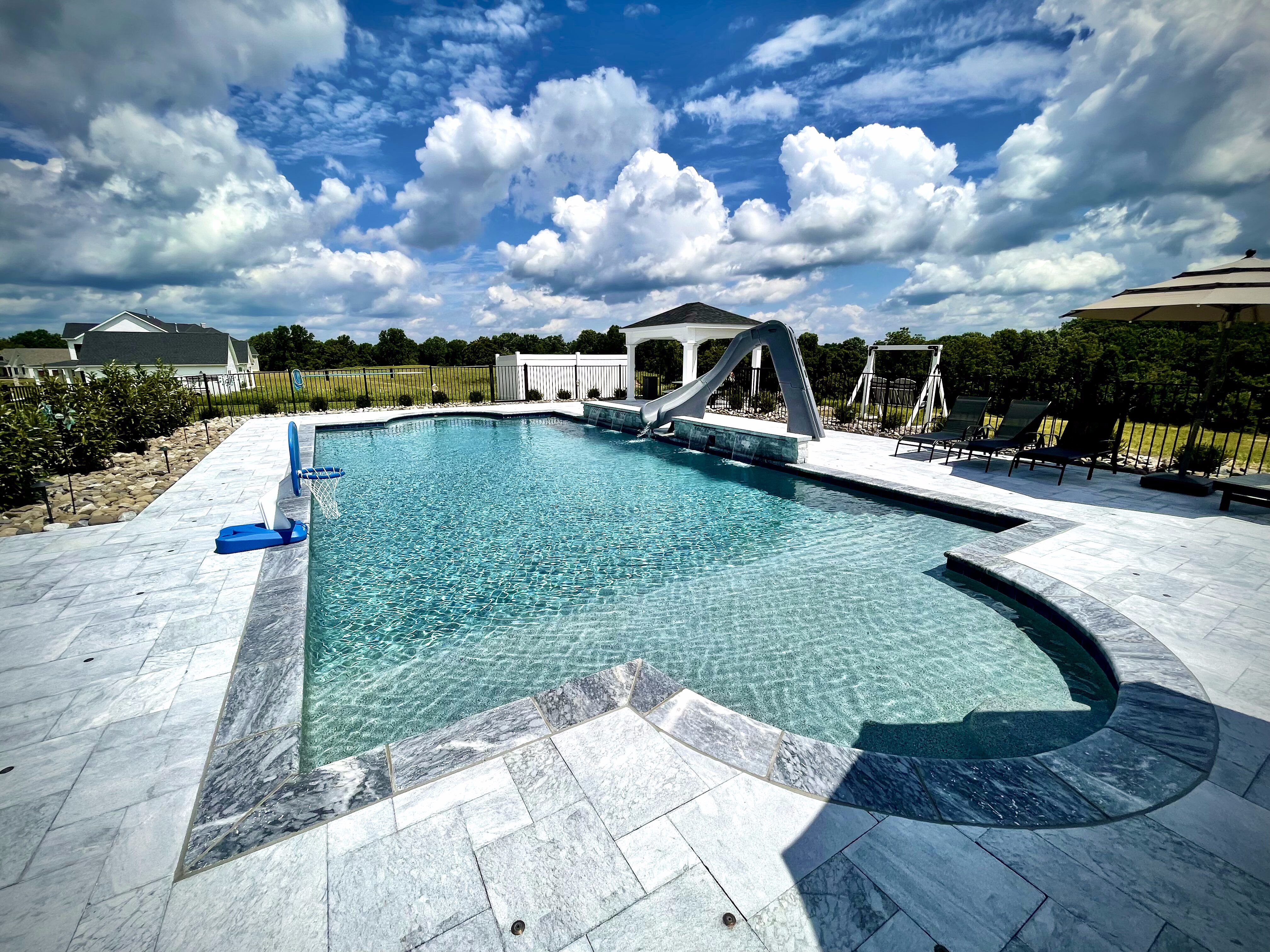 A sunny, wide-angle photo of a luxurious in-ground swimming pool with light blue water and a gray and white tiled deck.