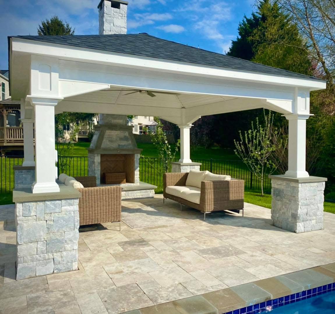 outdoor patio area with a white, covered pavilion. The pavilion has a dark roof and is supported by four white pillars. The base of the pillars is clad in light-colored stone. Under the pavilion, there is a stone fireplace and two wicker sofas with light-colored cushions. The patio floor is made of light-colored pavers, and the area is next to a swimming pool, visible in the foreground.