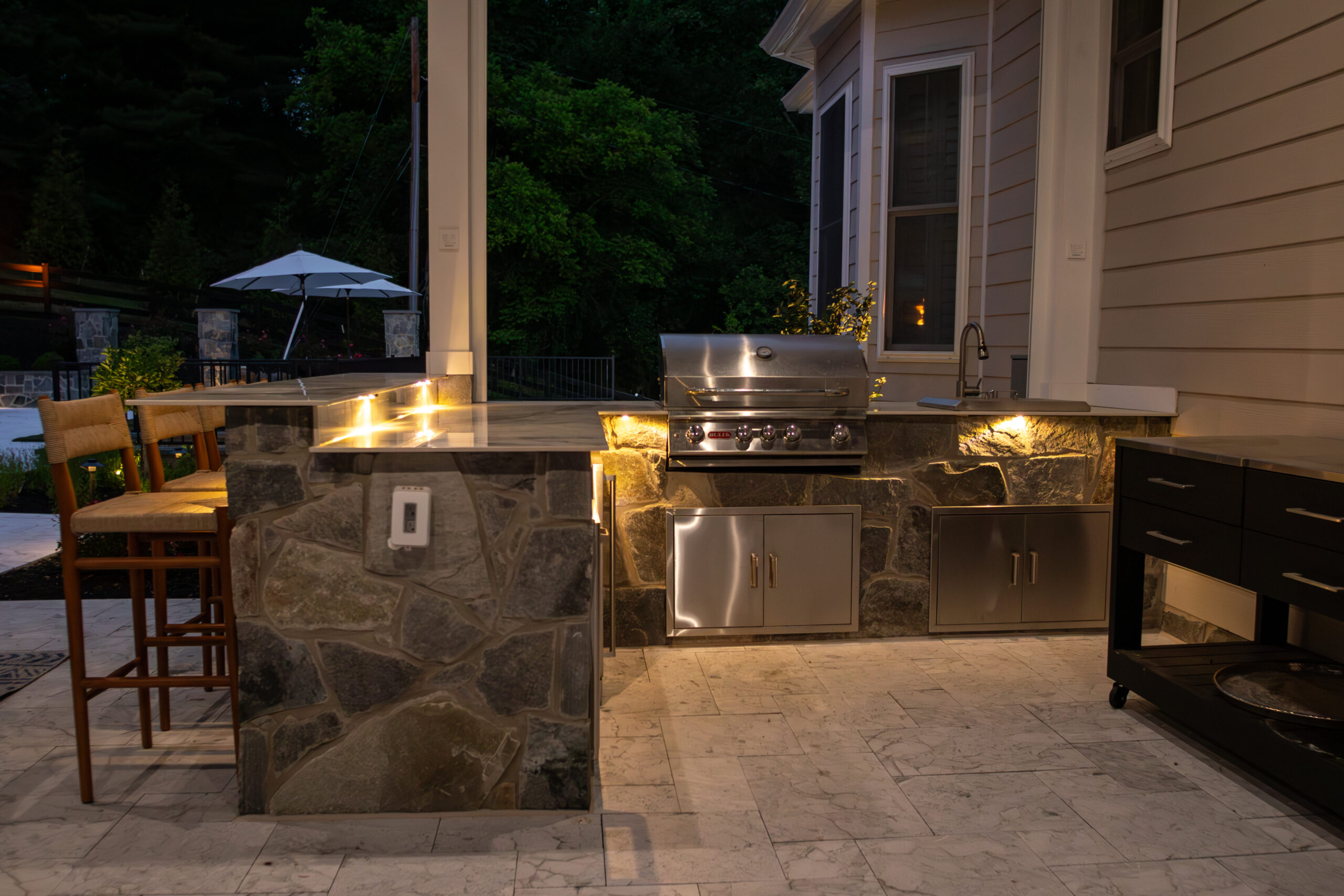 An image of an outdoor kitchen area at night, illuminated by soft lighting. The kitchen features a stone island with a stainless steel built-in grill and cabinets underneath.