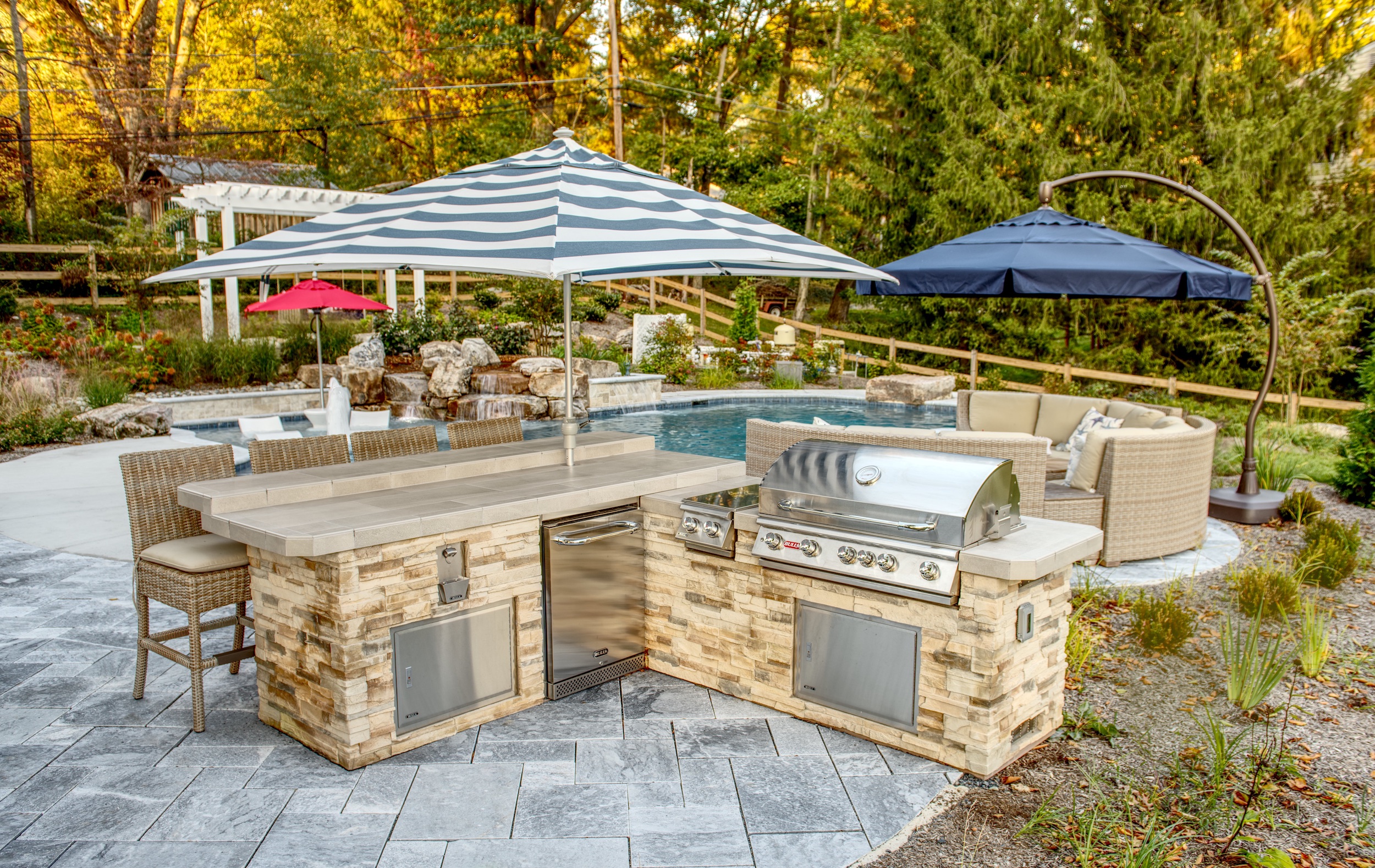 pscale outdoor living area featuring a custom L-shaped outdoor kitchen built with light brown stacked stone and a tan countertop. The kitchen includes a stainless steel grill, a compact refrigerator, and storage drawers.